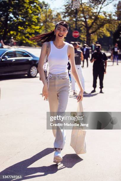 Chinese-American model Sherry Shi wears a fitted white tank top, gold earrings, a purple purse, tan pants with a shoelace belt, tan tote bag, and...