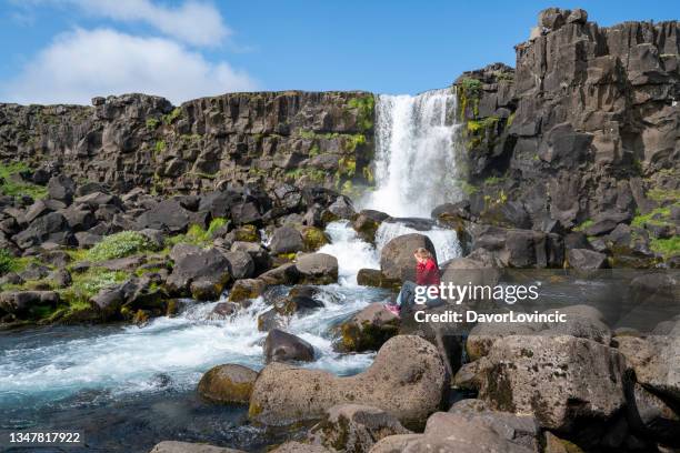 mature woman siting on rock and enjoying oxararfoss waterfall at mid-atlantic ridge, iceland - nationaal park pingvellir stockfoto's en -beelden