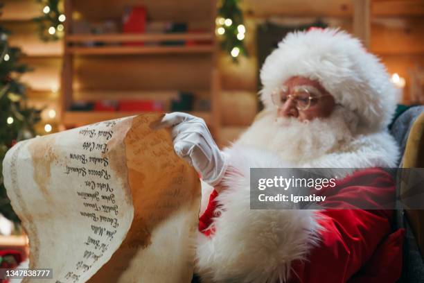 le père noël est assis sur une chaise avec un long rouleau de papier dans les mains - father christmas photos et images de collection