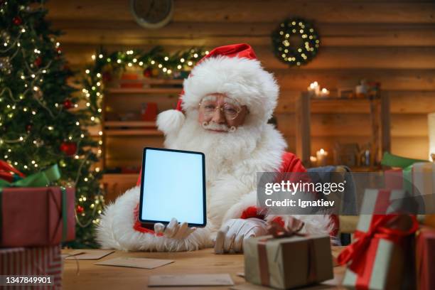 santa claus is sitting in a hut, with xmas decoration, showing the digital tablet in camera - retirement invitation stock pictures, royalty-free photos & images