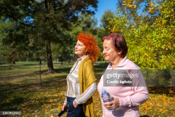 two senior women smiling while walking on path in forest - oranje haar stockfoto's en -beelden