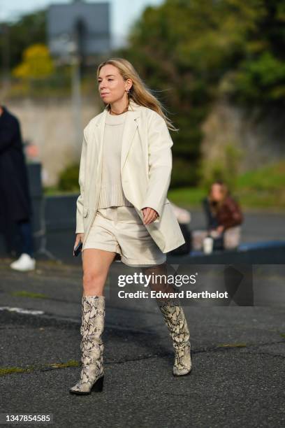 Guest wears gold earrings, a white latte wool pullover, a beige matte leather long blazer jacket, white latte denim shorts, silver rings, a gold...