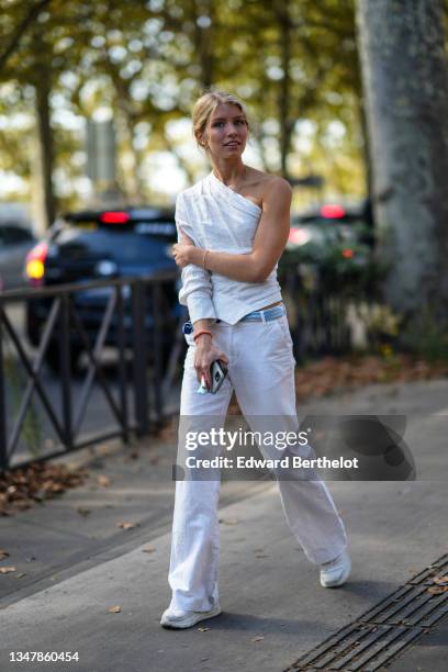 Guest wears gold earrings, a white cotton asymmetric sleeves t-shirt, a white and pale blue silk scarf as a belt, white flared pants, a pink and...