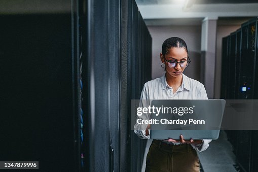 Shot of a young woman using a laptop while working in a server room Shot of a young woman using a laptop while working in a server room