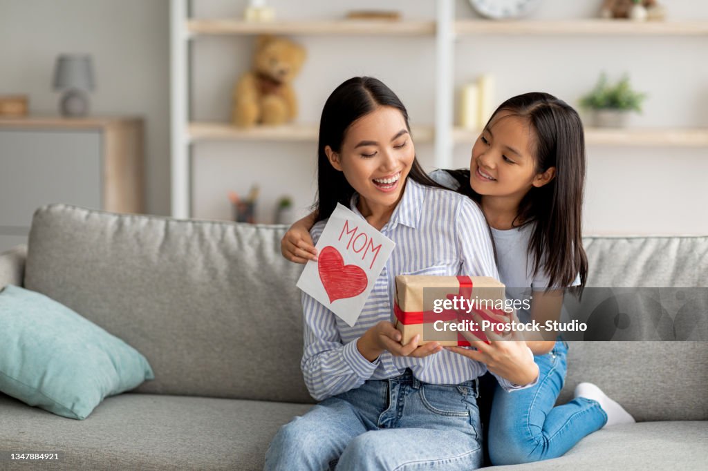 Congratulations. Loving asian daughter greeting surprised mom with Mother's Day, giving her handmade card and gift box