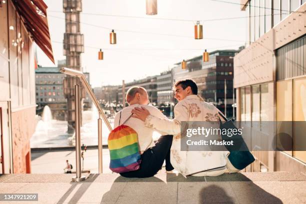 male friends talking while sitting on staircase in city during sunny day - stockholm summer stock-fotos und bilder