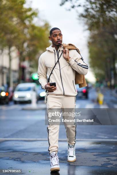 Jarrell Hargraves wears a beige suede jacket, a black woven leather bag, a watch, a white hoodie sportswear sweater, matching jogger pants, ankle...