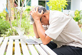 Portrait of an old elderly Asian man holds the head with his hand cause of stress after try to use a computer laptop in the backyard after retired. Concept of Ageism and Hobbies after retirement.