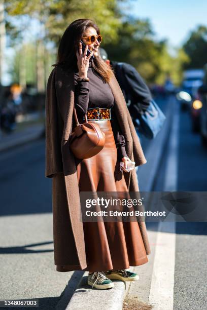 Guest wears brown sunglasses, rings, a dark brown turtleneck pullover, a brown long wool coat, a brown and orange tie and dye print pattern metallic...
