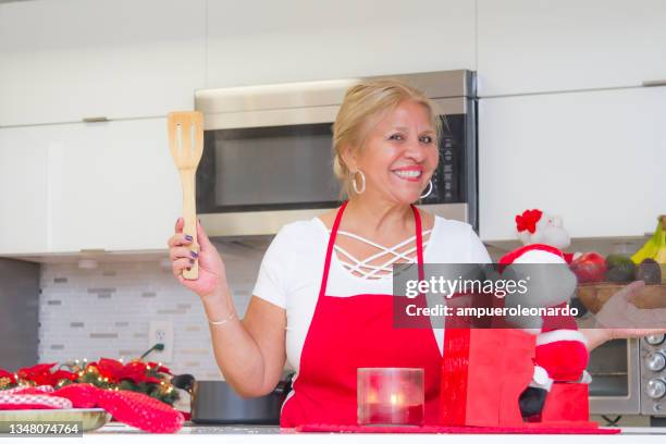 latin mature woman cooking christmas dinner / thanksgiving dinner for christmas night / thanksgiving day inside in a latin moderm home in miami, united states wearing white shirt and a red apron after corona virus covid-19 pandemic illness breakdown. - virtual thanksgiving stock pictures, royalty-free photos & images