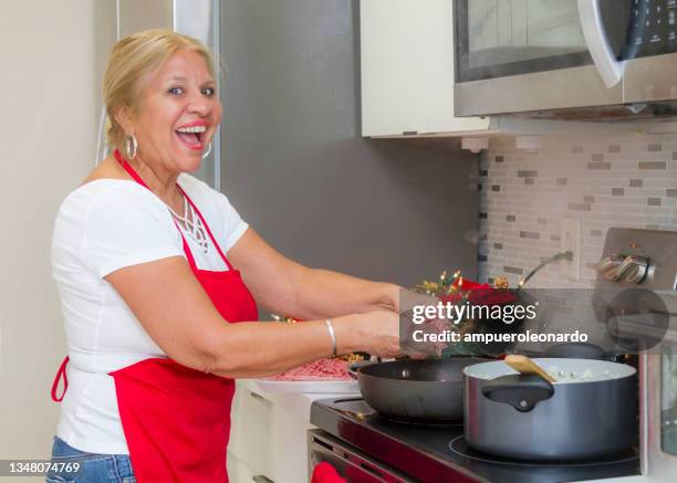 latin mature woman cooking christmas dinner / thanksgiving dinner for christmas night / thanksgiving day inside in a latin moderm home in miami, united states wearing white shirt and a red apron after corona virus covid-19 pandemic illness breakdown. - virtual thanksgiving stock pictures, royalty-free photos & images