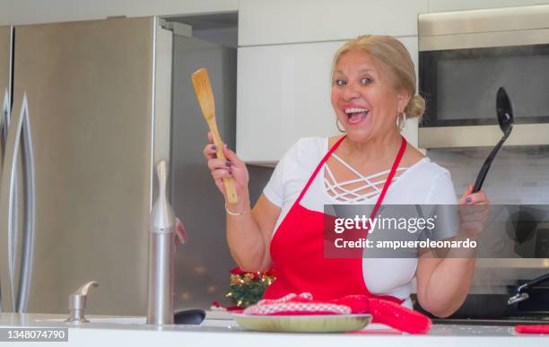 latin mature woman cooking christmas dinner / thanksgiving dinner for christmas night / thanksgiving day inside in a latin moderm home in miami, united states wearing white shirt and a red apron after corona virus covid-19 pandemic illness breakdown. - virtual thanksgiving stock pictures, royalty-free photos & images