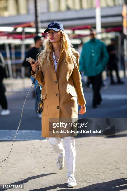 Guest wears navy blue cap from Ritz Paris, gold earrings form Chanel, a gold chain pendant necklace from Chanel, a white shirt, a beige wool coat,...