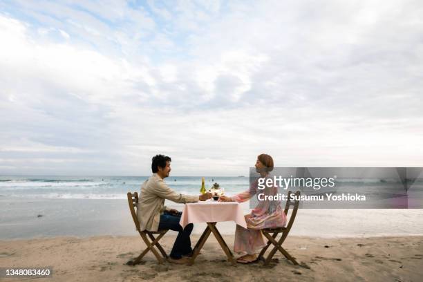 a couple enjoying a glass of wine while looking out at the ocean on a beautiful beach in the evening. - the japanese wife foto e immagini stock
