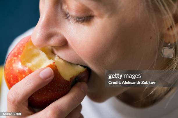 a red apple in the hands of a woman who is sitting on a bright-colored sofa at home. the girl eats a fruit, bites off a piece of juicy apple. cultivation of organic farm products. the concept of vegetarian, vegan, raw food and diet. - social bite stock pictures, royalty-free photos & images