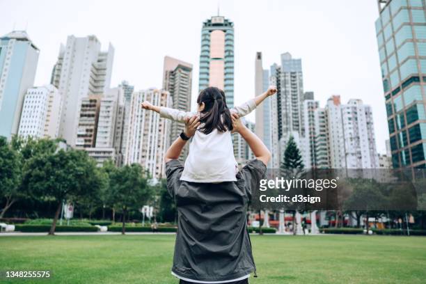 rear view of young asian father carrying happy little daughter on shoulders in urban park. enjoying father-daughter bonding time together. family love and lifestyle - carrying on shoulders stock pictures, royalty-free photos & images