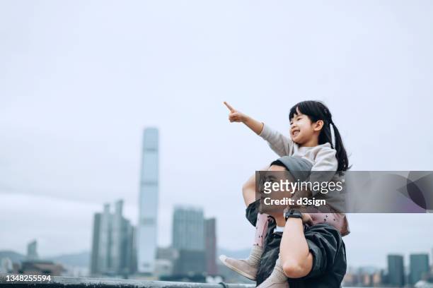 young asian father giving little daughter piggyback ride by the promenade of victoria harbour, little girl pointing far away while looking over the beautiful city skyline - apuntar fotografías e imágenes de stock