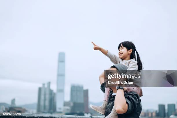 young asian father giving little daughter piggyback ride by the promenade of victoria harbour, little girl pointing far away while looking over the beautiful city skyline - looking to the future photos et images de collection