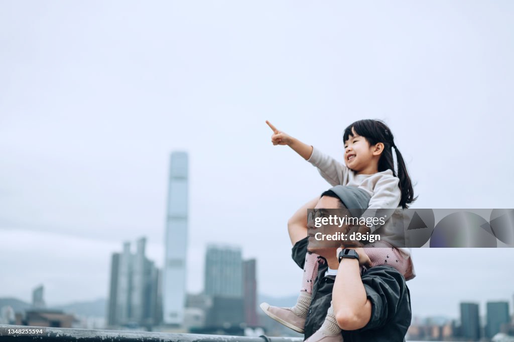 Young Asian father giving little daughter piggyback ride by the promenade of Victoria harbour, little girl pointing far away while looking over the beautiful city skyline