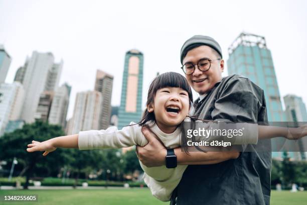 happy little asian girl having fun with her father outdoors in urban park. she is pretending to be an airplane, flying up in the sky while her father lifting her into the air. family love and lifestyle - only kids at sky stockfoto's en -beelden