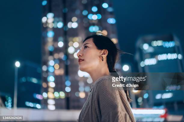 low angle portrait of young asian businesswoman standing in downtown city street, looking up with confidence against illuminated urban commercial buildings and vibrant city street lights at night. business on the go - night before fotografías e imágenes de stock