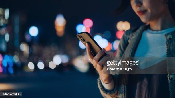close up of young asian woman using mobile app on smartphone to arrange taxi ride in downtown city street at night, with illuminated city scene as background - application modernization stock pictures, royalty-free photos & images