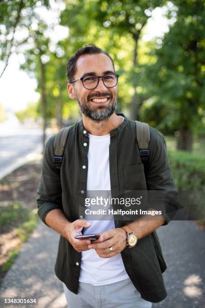 portrait of happy mature man walking and using smartphone outdoors in park. - casual man walking stock pictures, royalty-free photos & images