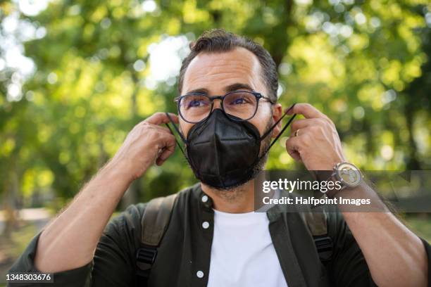 portrait of mature man walking and putting on face mask outdoors in park. - n95 ademhalingsmasker stockfoto's en -beelden