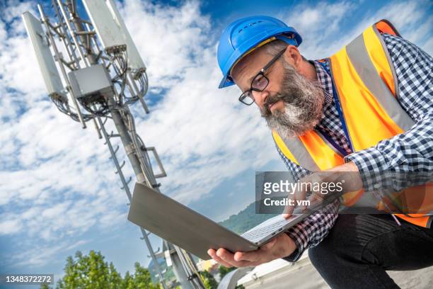 bearded technician in reflective vest working on laptop computer outdoors - communication tower stock pictures, royalty-free photos & images