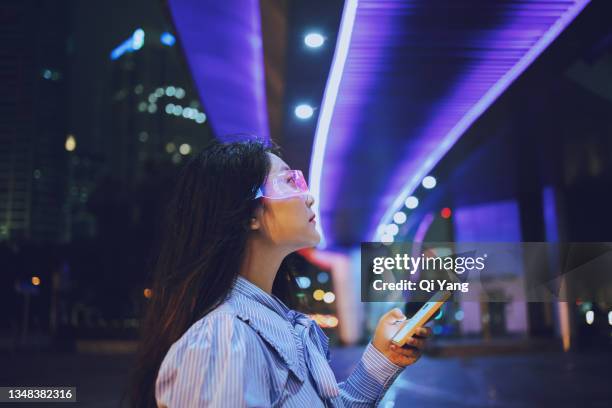 young asian woman using mobile phone while standing under a pedestrian bridge glowing at night - innovatie stockfoto's en -beelden