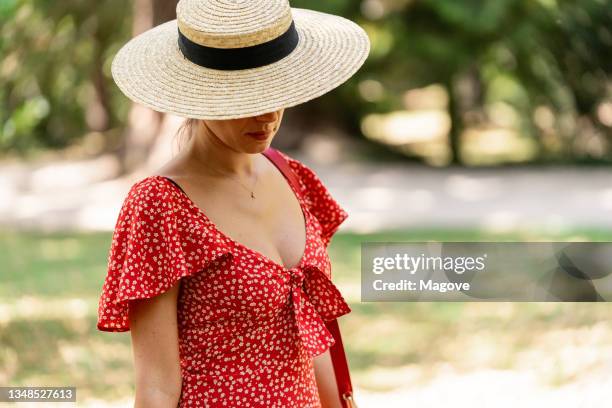 woman in a red dress and straw hat standing outdoors in a park. - strohoed stockfoto's en -beelden