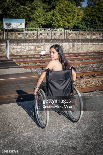 disabled young woman sitting in a wheelchair in front of the train tracks while looking frustrated at the camera facing the difficulties of moving in the city. - paraplégico imagens e fotografias de stock