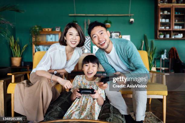 happy young asian family and daughter spending some quality time together, having fun playing video games while sitting on the sofa in the living room at home - family teenager home life stock pictures, royalty-free photos & images