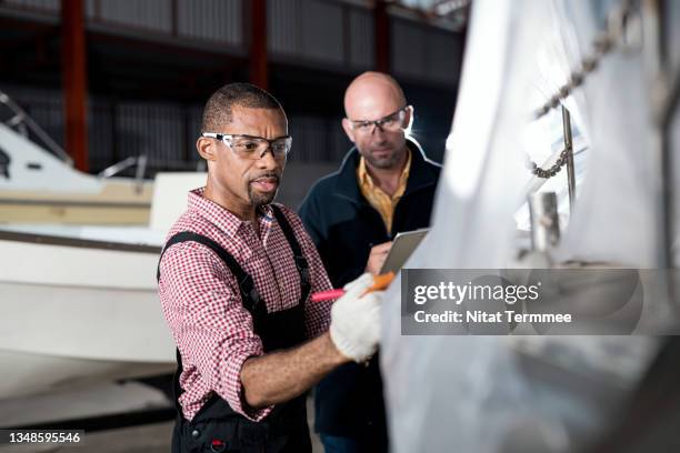 measuring coating quality, cleaning, and painting a boat’s hull. african american senior boat mechanic and customer examine crack a fiberglass outer surface (body of a vessel) to restoring and renovation in a boathouse. - carrosserie stockfoto's en -beelden