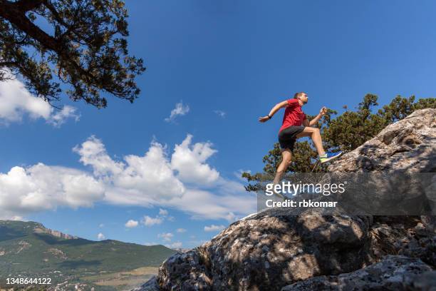 young fitness man in red sports shirt and shorts running up the edge of a rocky mountain to the top - leap forward stock pictures, royalty-free photos & images