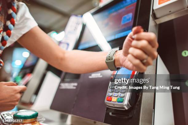 a female shopper making contactless payment using smart watch at self-service checkout in supermarket - watch payment stock pictures, royalty-free photos & images