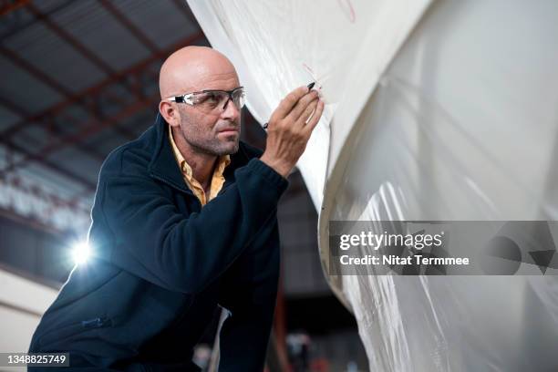 low angle of a male boat owner used a marker pen to check on fiberglass surface outer of v-shaped hulls for surface restoration, prepare painting, and anti-rust coating in his boathouse. marine paint, coatings, and boat renovation business. - vetroresina foto e immagini stock