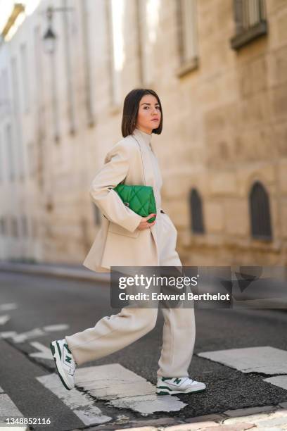 Alexandra Pereira wears silver earrings, a beige turtleneck pullover, matching beige wool large pants, a beige blazer jacket, a green shiny leather...