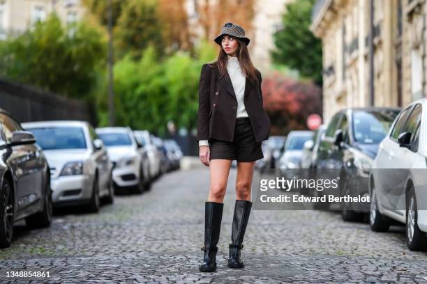 Amanda Derhy wears a brown and beige houndstooth print pattern bob / hat, a beige ribbed wool turtleneck pullover with gold buttons on the neck, a...