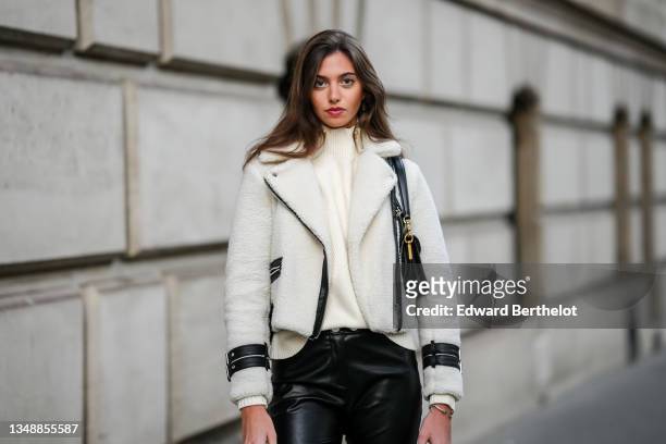 Amanda Derhy wears gold earrings, a beige ribbed wool turtleneck pullover with gold buttons on the neck, white sheep with black leather yoke aviator...