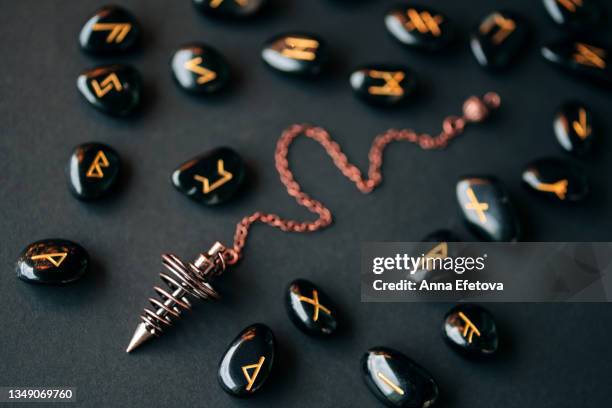 bronze dowsing pendulum and many runes made of obsidian with gold text arranged on black background. concept of magic esoteric rituals. macro photography in flat lay style - rune symbols stock pictures, royalty-free photos & images