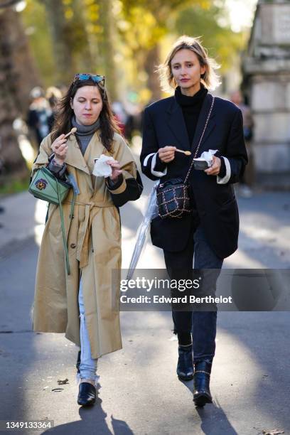 Guest wears sunglasses, a gray wool ribbed pullover, a beige long belted trench coat, a green leather with purple and yellow paint print pattern...