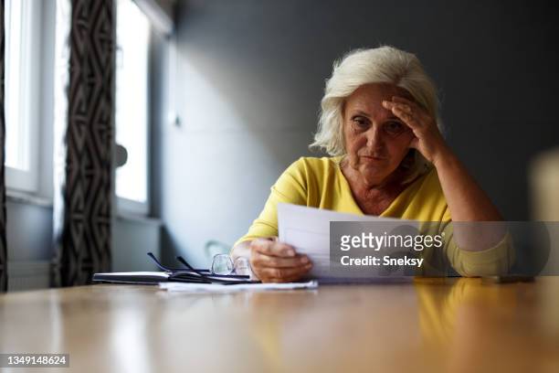 senior woman is sitting at the table, holding her forehead and reading reports. - unhappy woman blonde glasses stock pictures, royalty-free photos & images