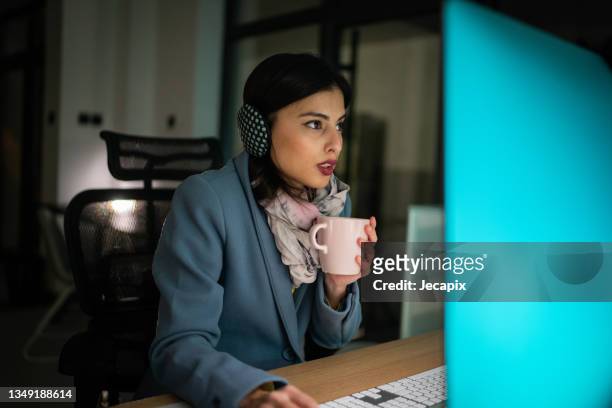 femme en vêtements chauds assise dans un bureau glacial et travaillant sur un ordinateur avec une tasse de café chaude à la main - froid photos et images de collection