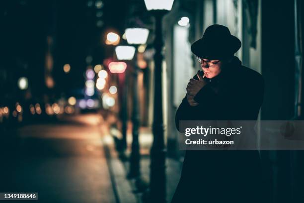 gutaussehender mann mit hut, der eine zigarette auf der stadtstraße anzünden - film noir style stock-fotos und bilder