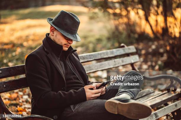 gutaussehender mann mit hut, der nachrichten am telefon im park überprüft - cold war spy stock-fotos und bilder