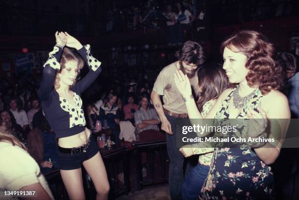 Female clubbers, one wearing black hotpants with a cropped blue top with polka dot detail, the other wearing a floral pattern dress, the crowded...