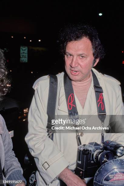 America photographer Ron Galella with a Nikon camera hanging from a strap around his neck, standing beside a parking meter, February 1977.