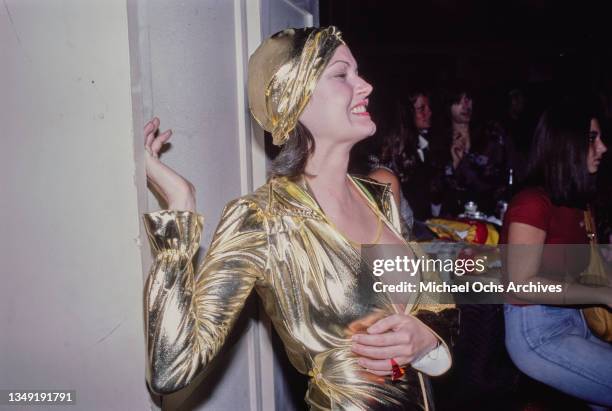 Woman wearing a gold lame outfit with a plunging neckline and a matching turban, leaning against a white wall, September 1976.