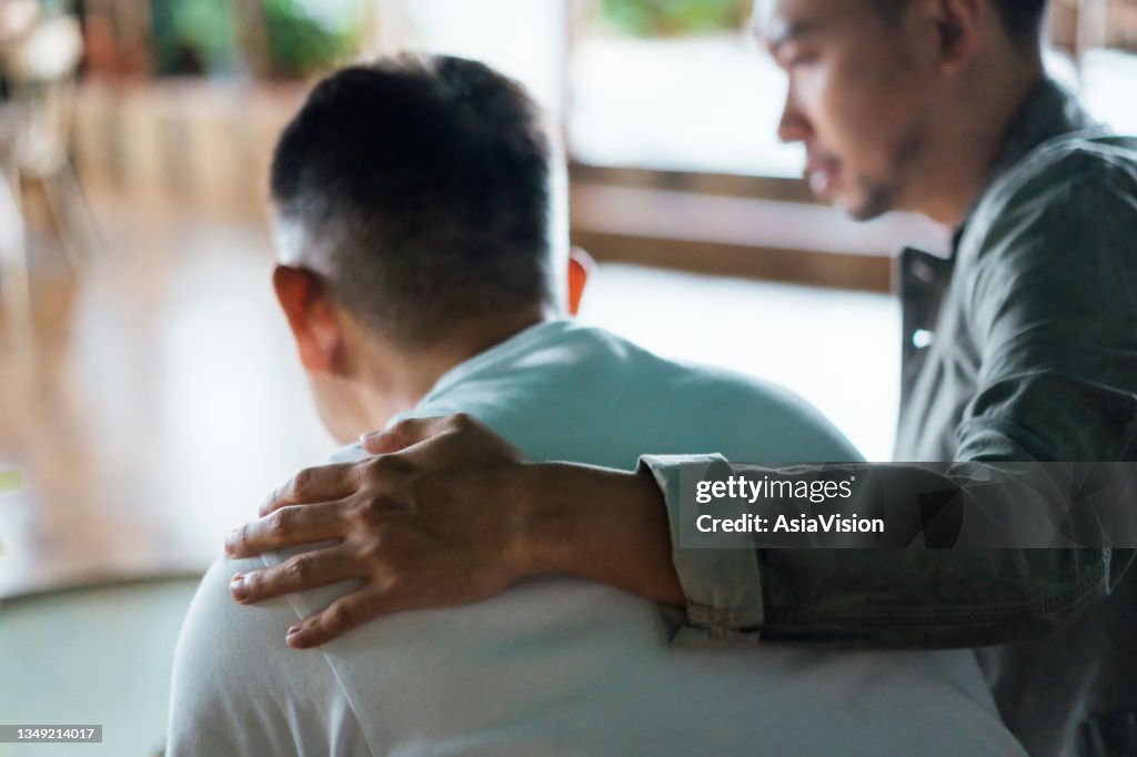 Rear view of son and elderly father sitting together at home. Son caring for his father, putting hand on his shoulder, comforting and consoling him. Family love, bonding, care and confidence